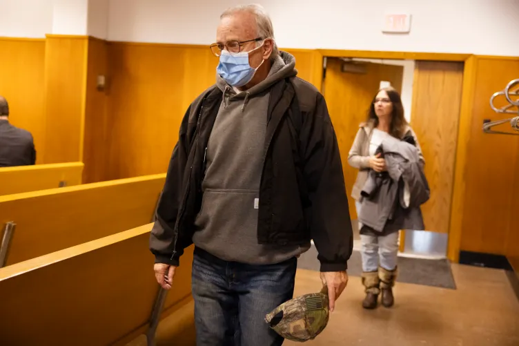 A man wearing a face mask, glasses and a jacket over a sweatshirt enters a courtroom while carrying a camouflage baseball hat.