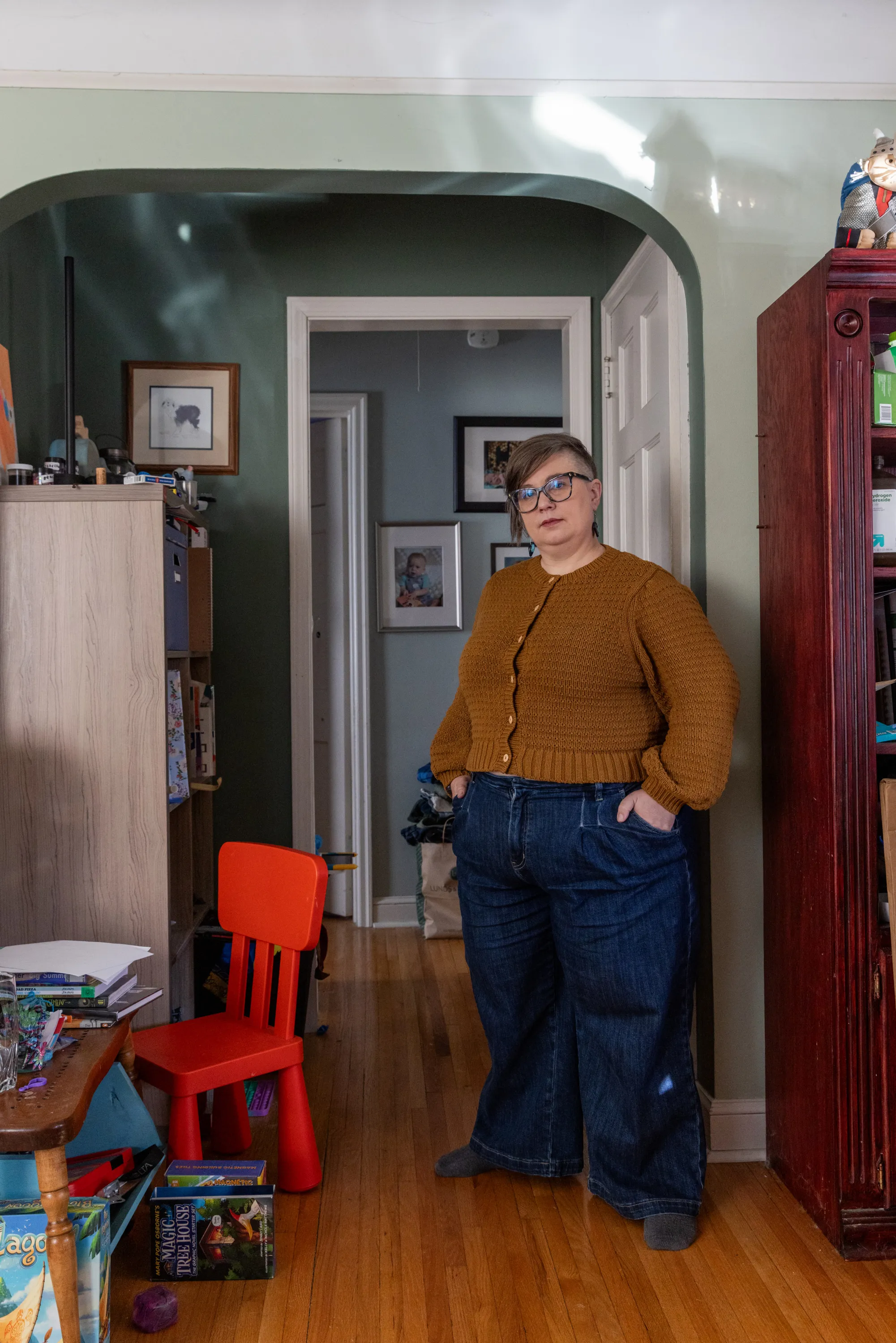 A woman stands with her hands in her pockets, looking at the camera, in the living room of her home. Next to her is a small table and small red chair for a child, with toys and books on the table and the floor. Behind her are family photos on the wall.