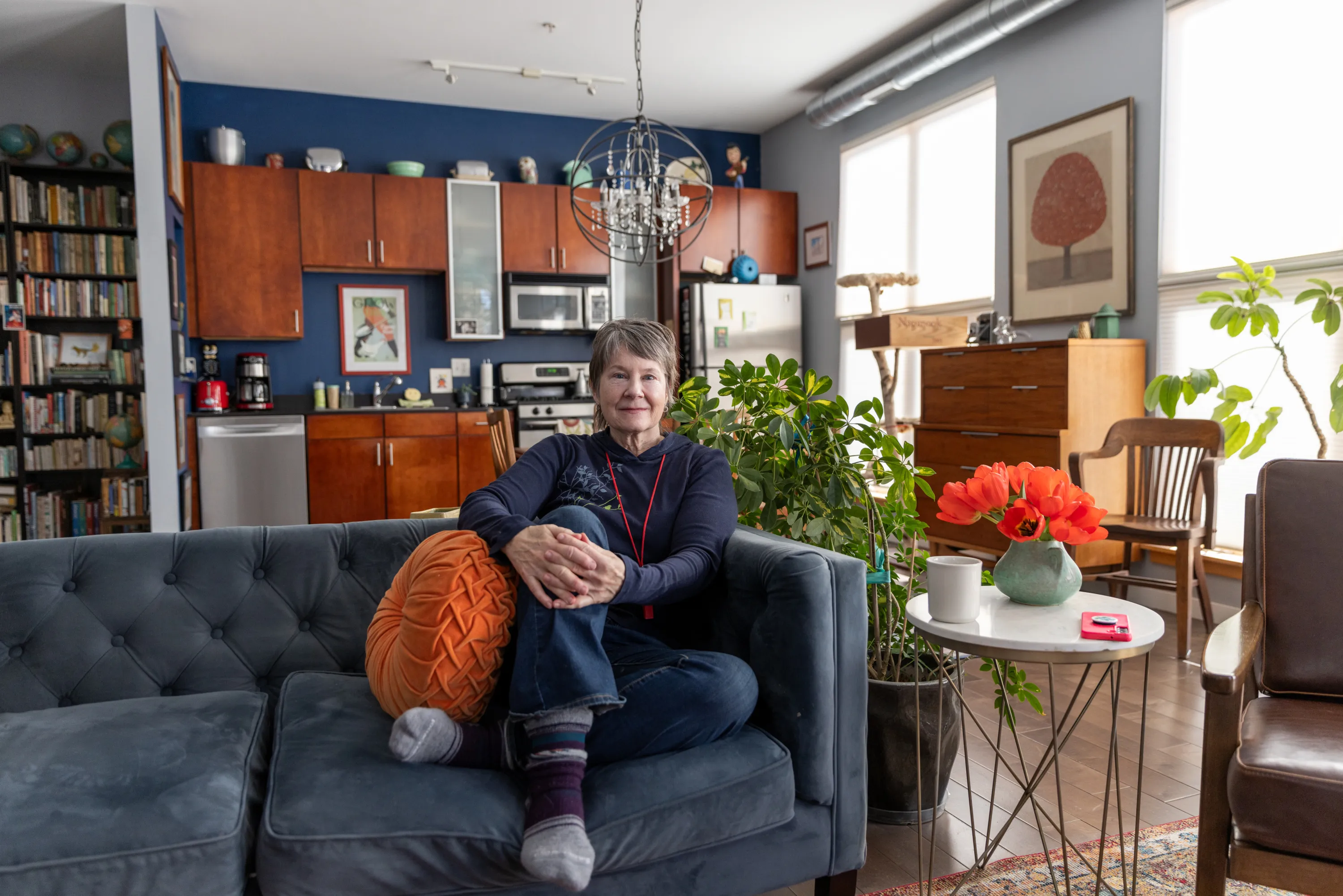 A woman sits with her hands clasped over her leg, looking at the camera. She is on a sofa in the living room of a condo, with several houseplants, paintings on the walls, a full bookshelf and her kitchen in the background.