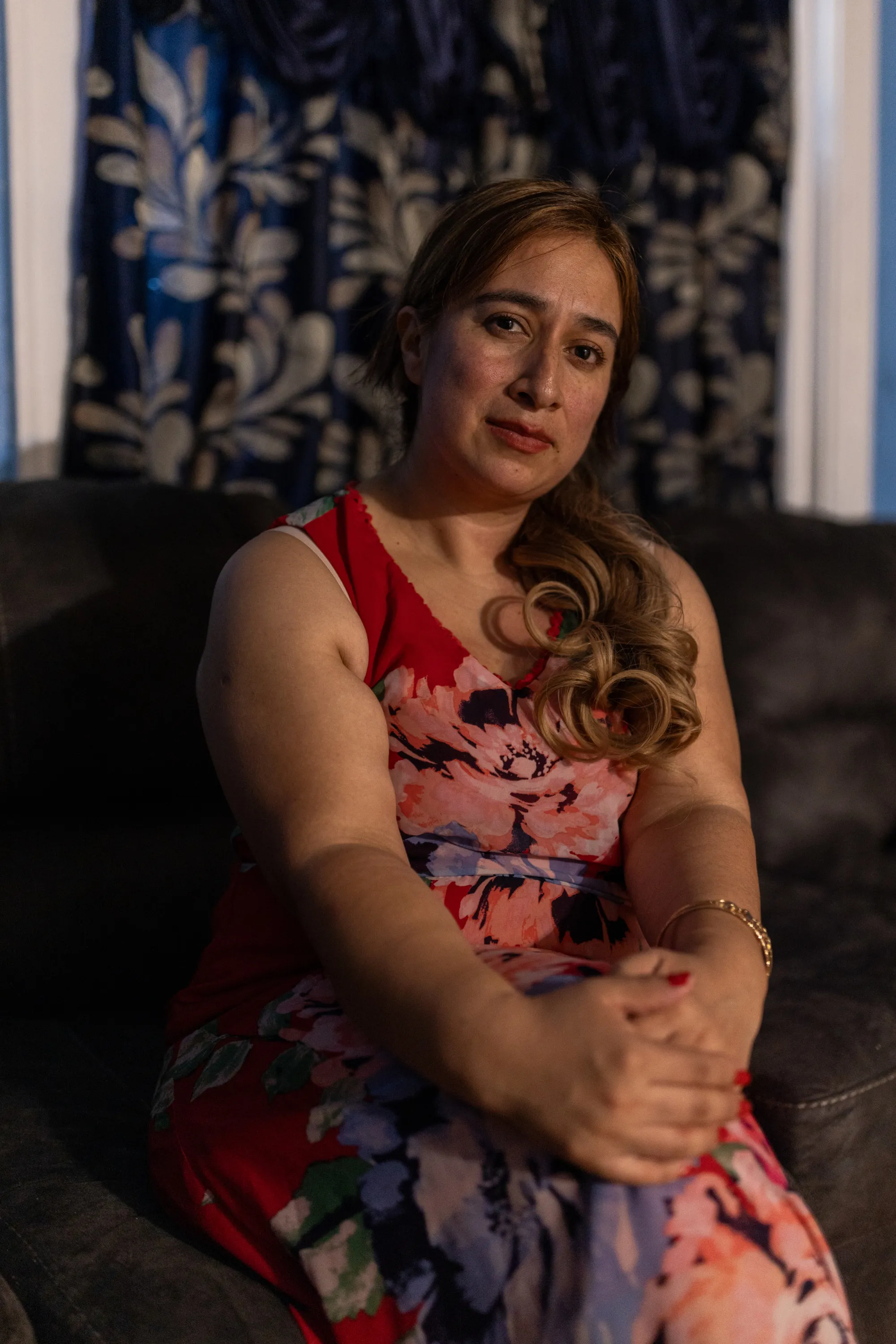 A woman sits in a dark room, looking at the camera, wearing a red floral print dress.
