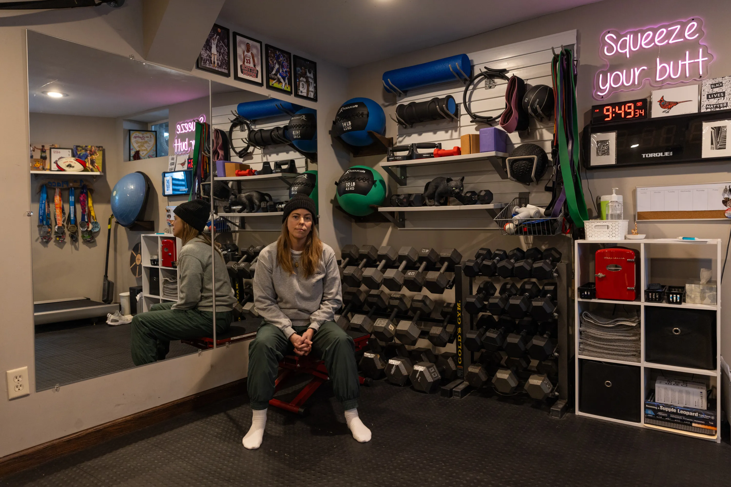 A woman sits in a basement gym, wearing a sweatshirt and athletic pants. She is reflected in a mirror on the wall behind her. There are lines of weights and other gym equipment and a neon sign that says “squeeze your butt.”