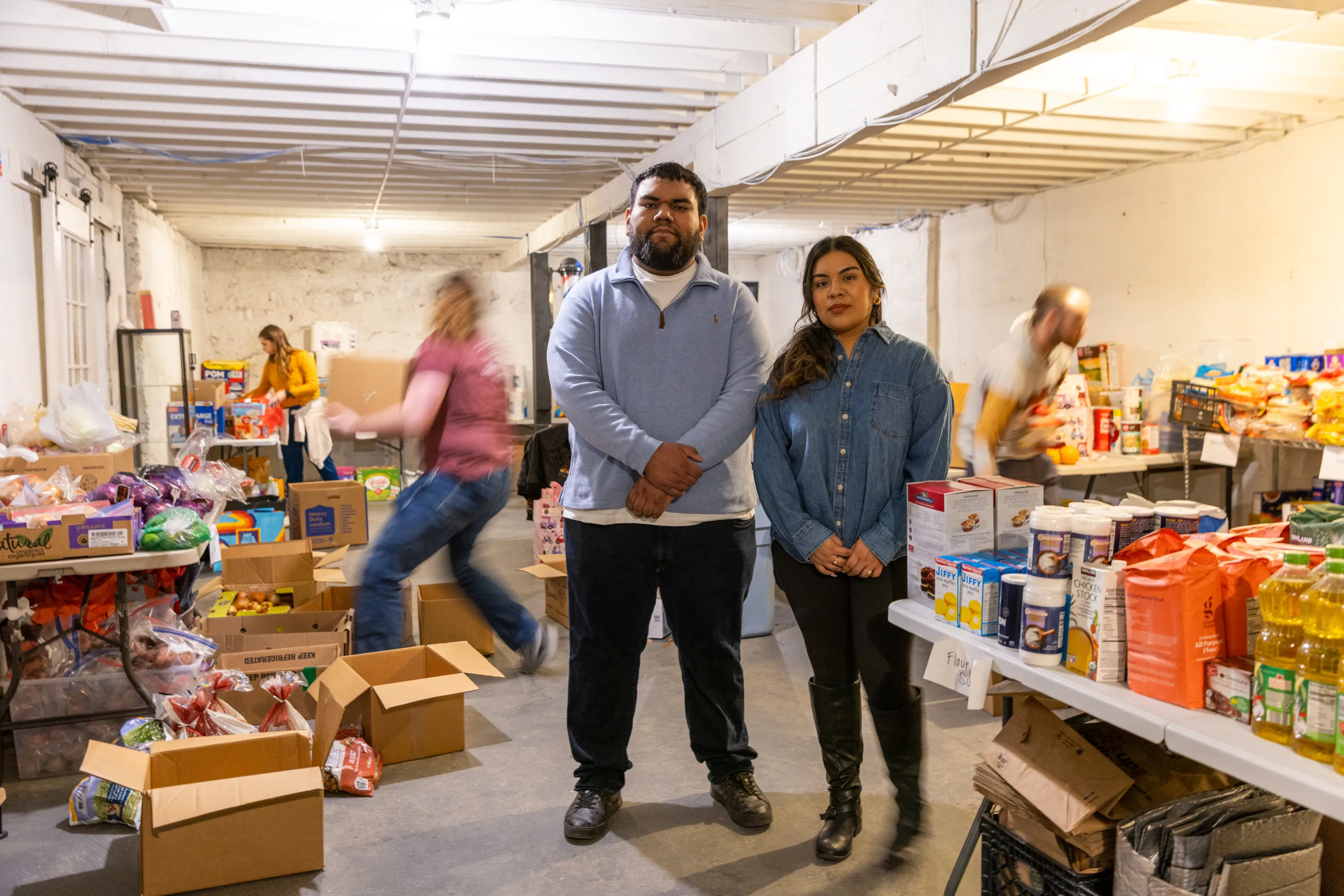 A man and woman stand together in a basement, looking at the camera. Around them are stacks of food, in cardboard boxes on the floor or on tables, and the blurred movement of several people moving and arranging the food.