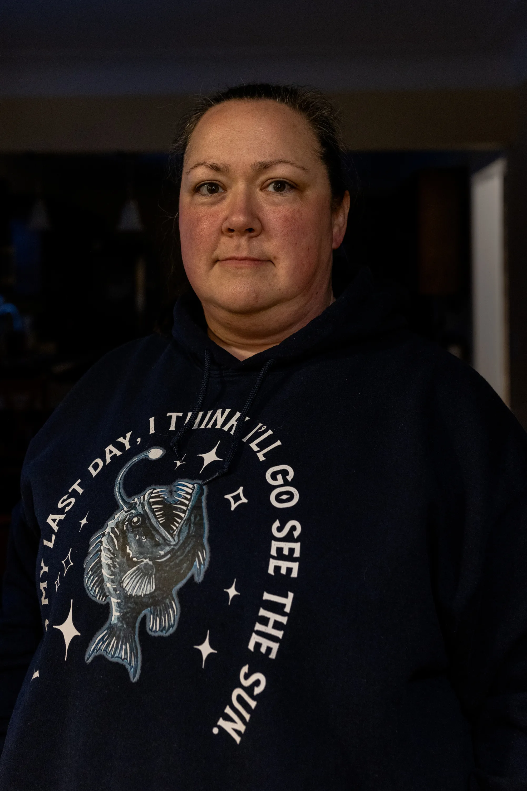 A woman in a dark room looks at the camera. She wears a sweatshirt with an image of an anglerfish and the words “my last day, I think I’ll go see the sun.”