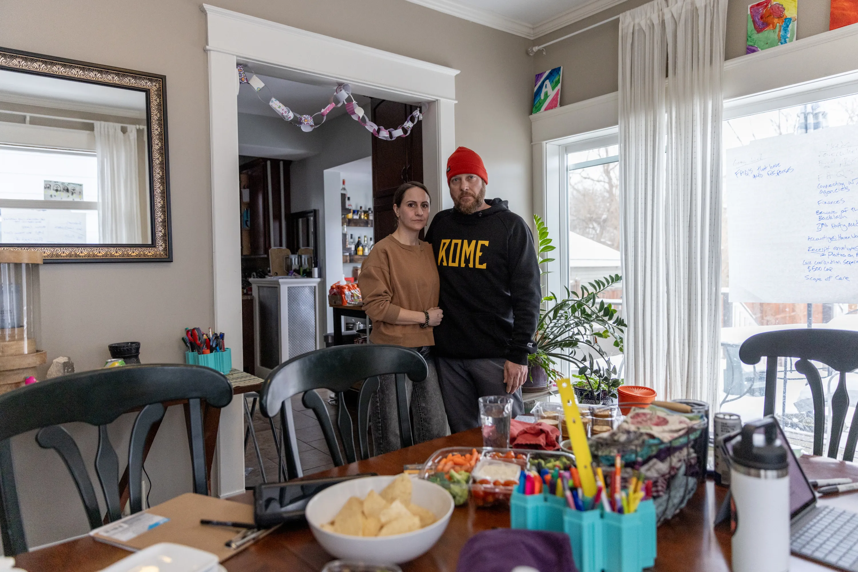 A woman and a man stand together in a dining room. In front of them, a dining room table is covered with snacks, drinks, a pencil holder full of pencils, a laptop and a tablet. On a window next to them, a large sheet of paper is taped up with meeting notes written on it.