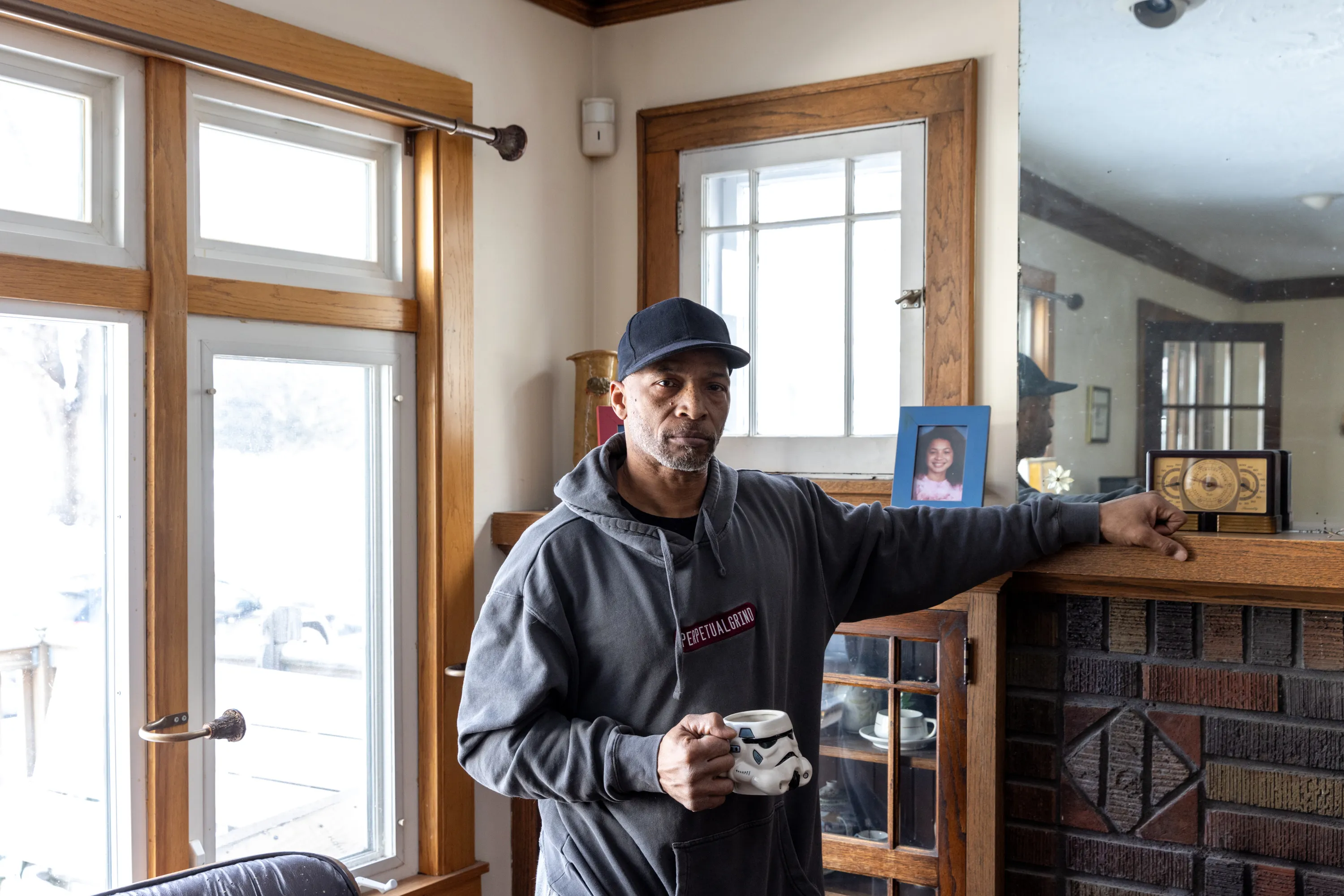 A man stands looking at the camera in his living room, with a mirror and a framed photo of a young girl in the background. He wears a sweatshirt that says “perpetual grind” and holds a Star Wars Stormtrooper coffee mug.