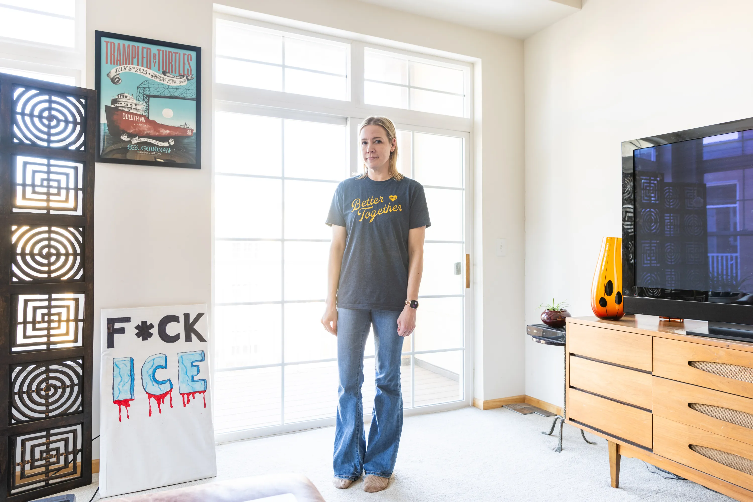 A woman stands in a bright condo, looking at the camera. A poster that says “Trampled by Turtles” is framed on the wall, and resting against the wall is a protest sign that reads “F*ck ICE.”
