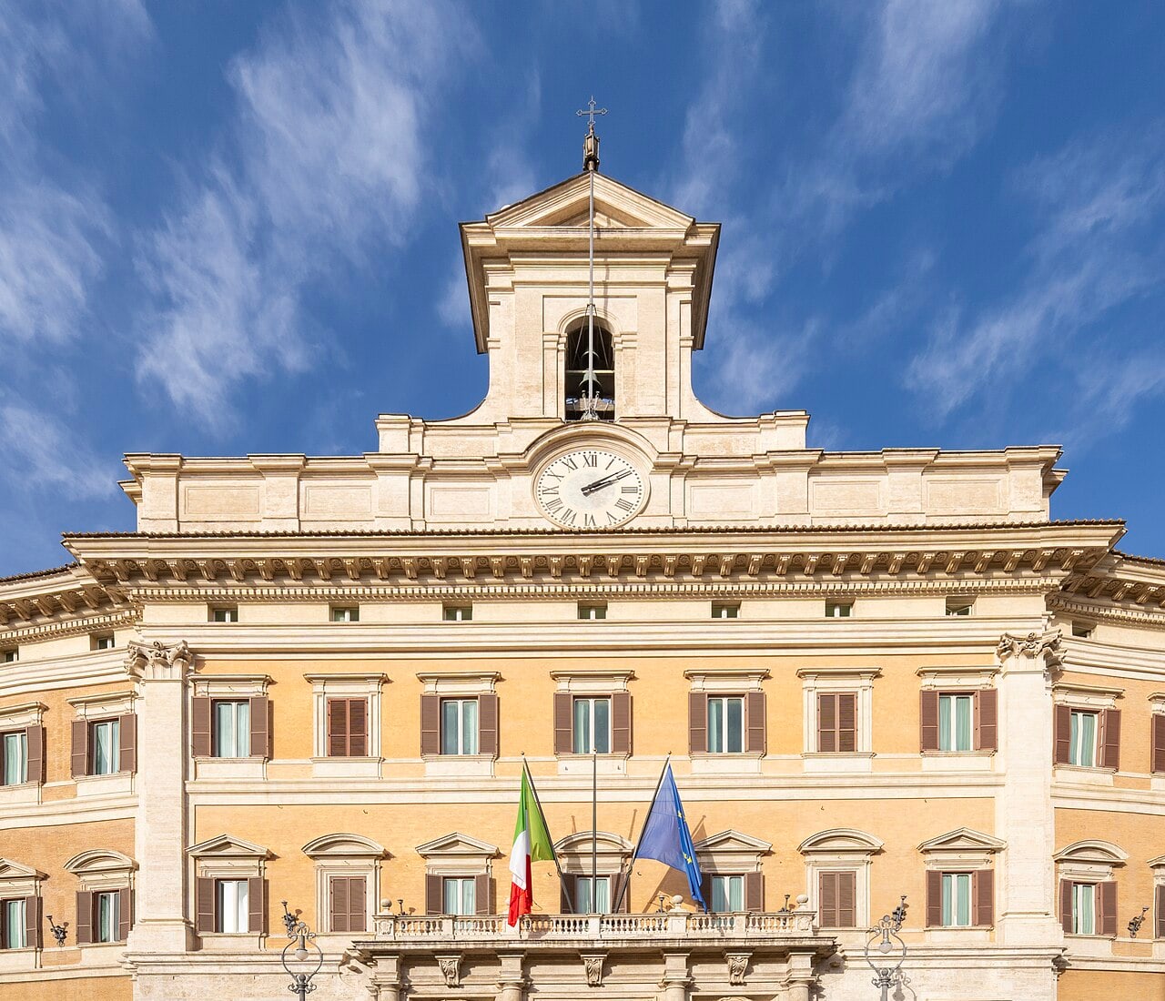La facciata di Palazzo Montecitorio a Roma sotto un cielo azzurro, con il campanile e l'orologio in alto e le bandiere italiana ed europea sul balcone