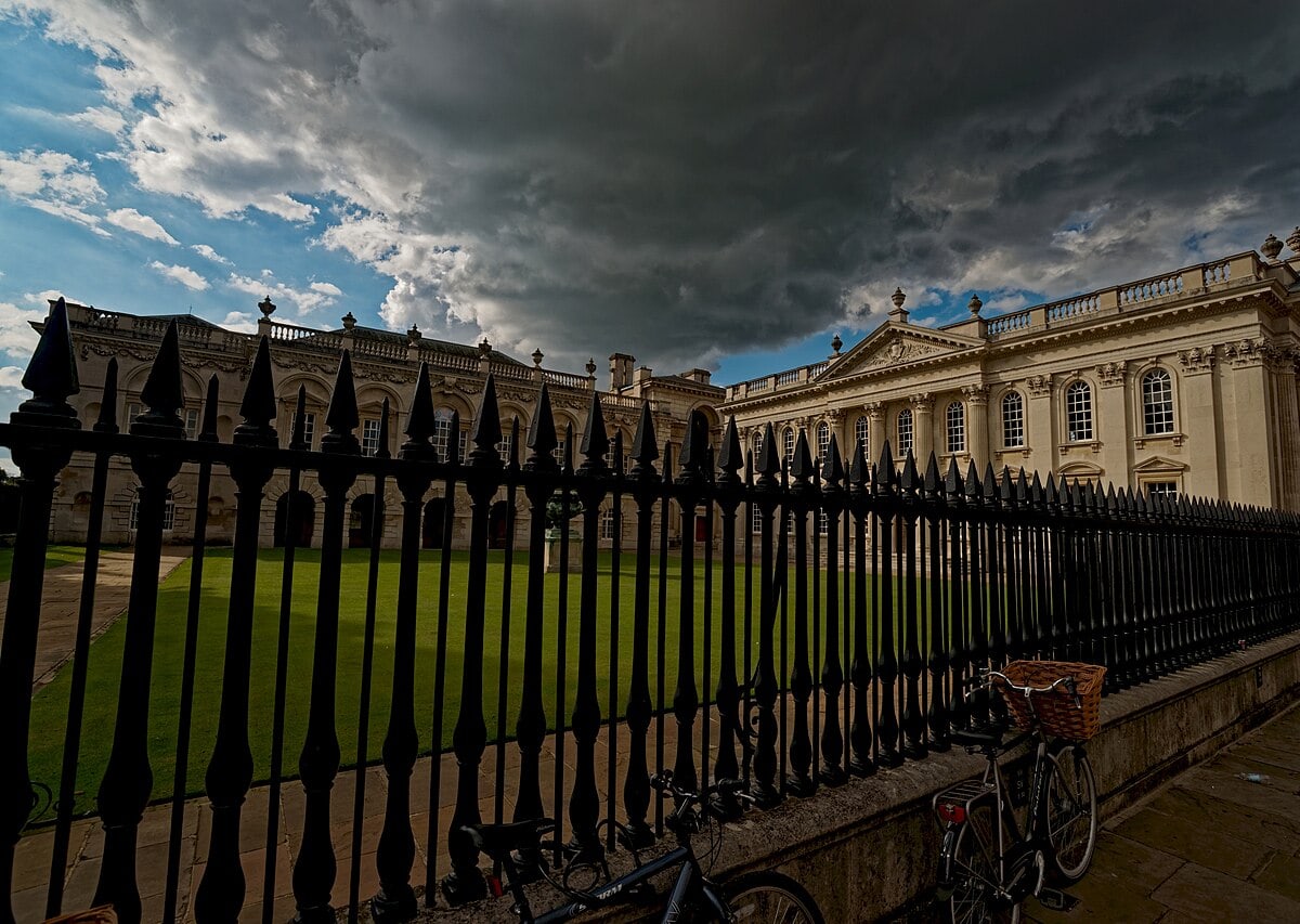 La Senate House dell'Università di Cambridge, in pietra chiara con colonne e balaustra, vista da King's Parade oltre una cancellata nera, con il prato davanti e un cielo carico di nuvole