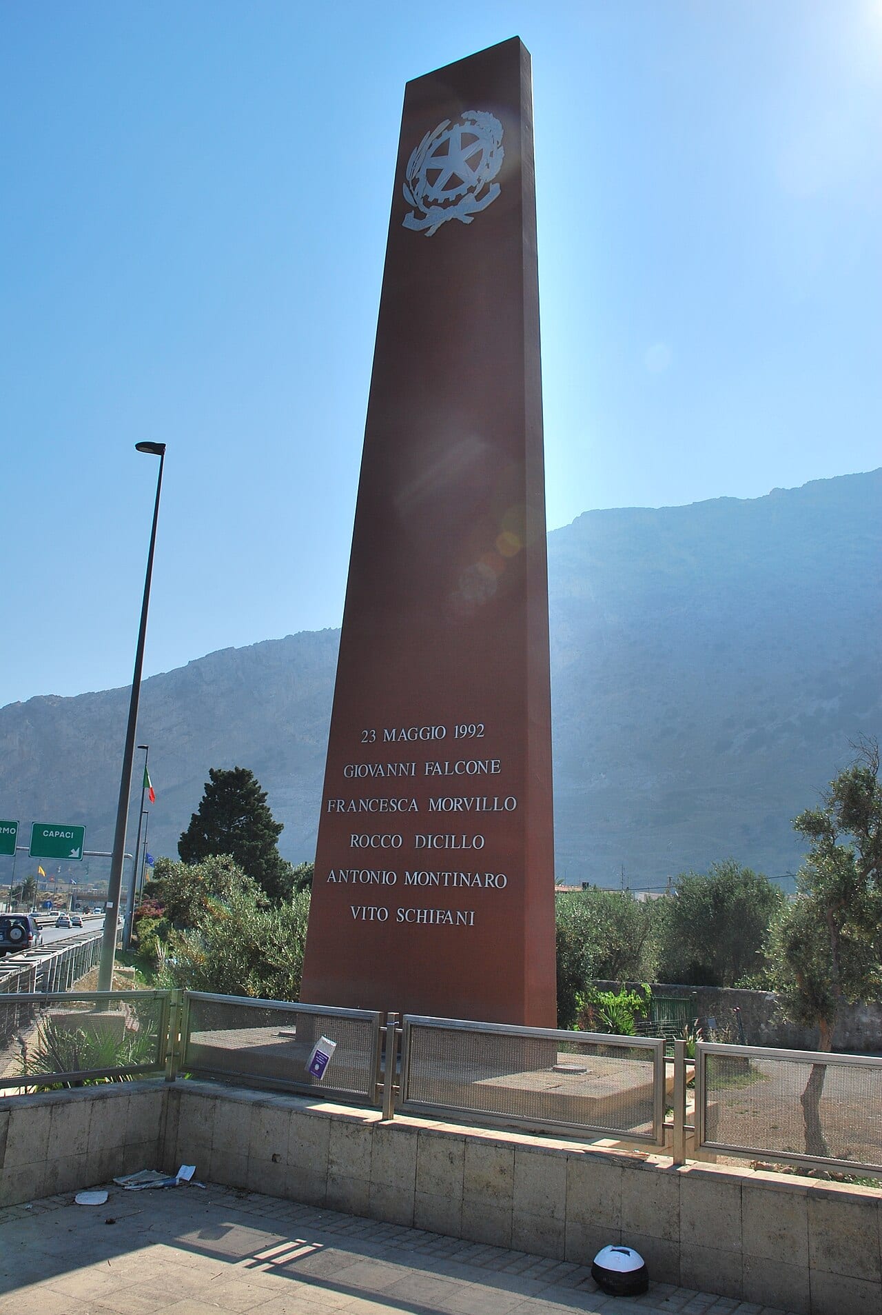 Tall red rectangular stele with the emblem of the Republic at the top and, below, the inscription "23 maggio 1992" and five names; a rocky mountain behind, the motorway junction with a "Capaci" sign on the left