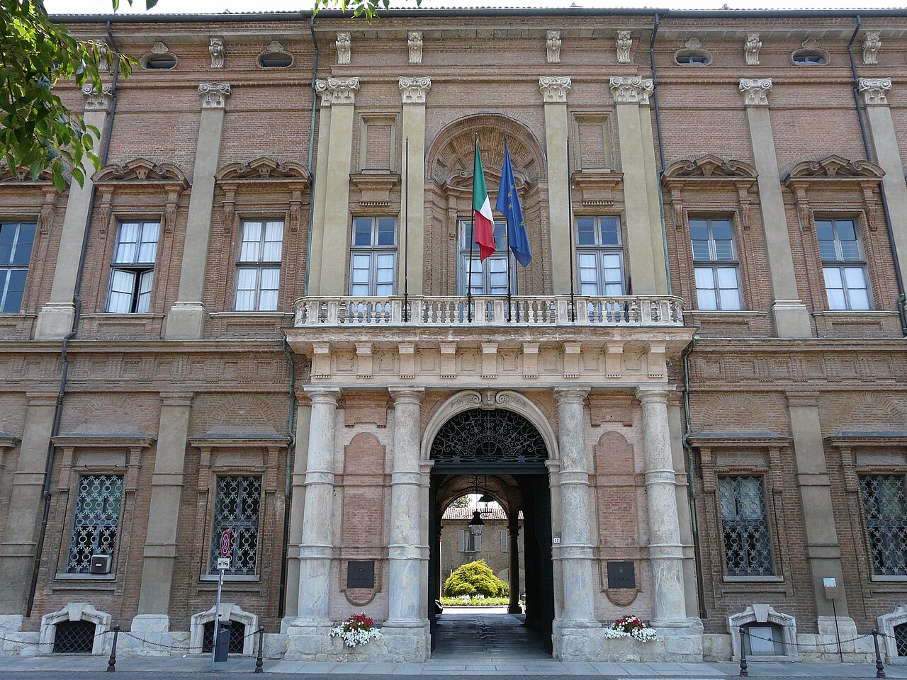 La facciata in mattoni di Palazzo Ghilini ad Alessandria, con il portone ad arco, il balcone con le bandiere italiana ed europea e le finestre con le grate